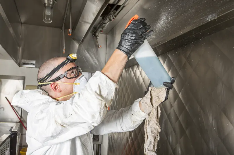 Technician in white coveralls, goggles and a headlamp reaching overhead with a spray bottle and cloth to wipe the underside of a stainless steel exhaust canopy, photographed from below