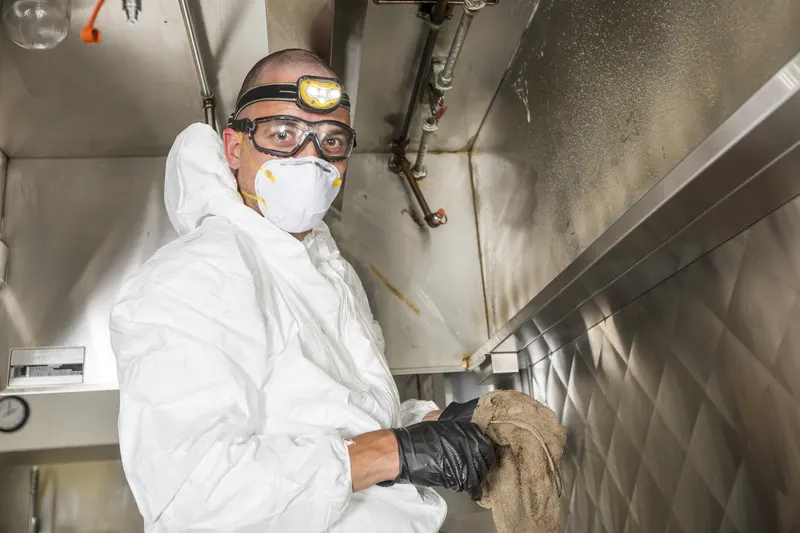 Technician in a white coverall, goggles and headlamp holding a cloth at the filter slot along the front rail of a kitchen exhaust hood